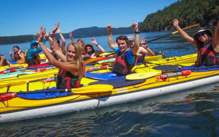 a group of students in kayaks raise their hands in celebration on an outward bound course